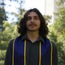 Man with shoulder length brown hair and facial hair wearing a graduation sash