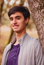 Man in a purple shirt and gray sweather smiles in front of a tree