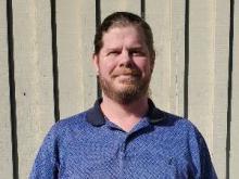 Man with a beard smiles in front of a gray fence